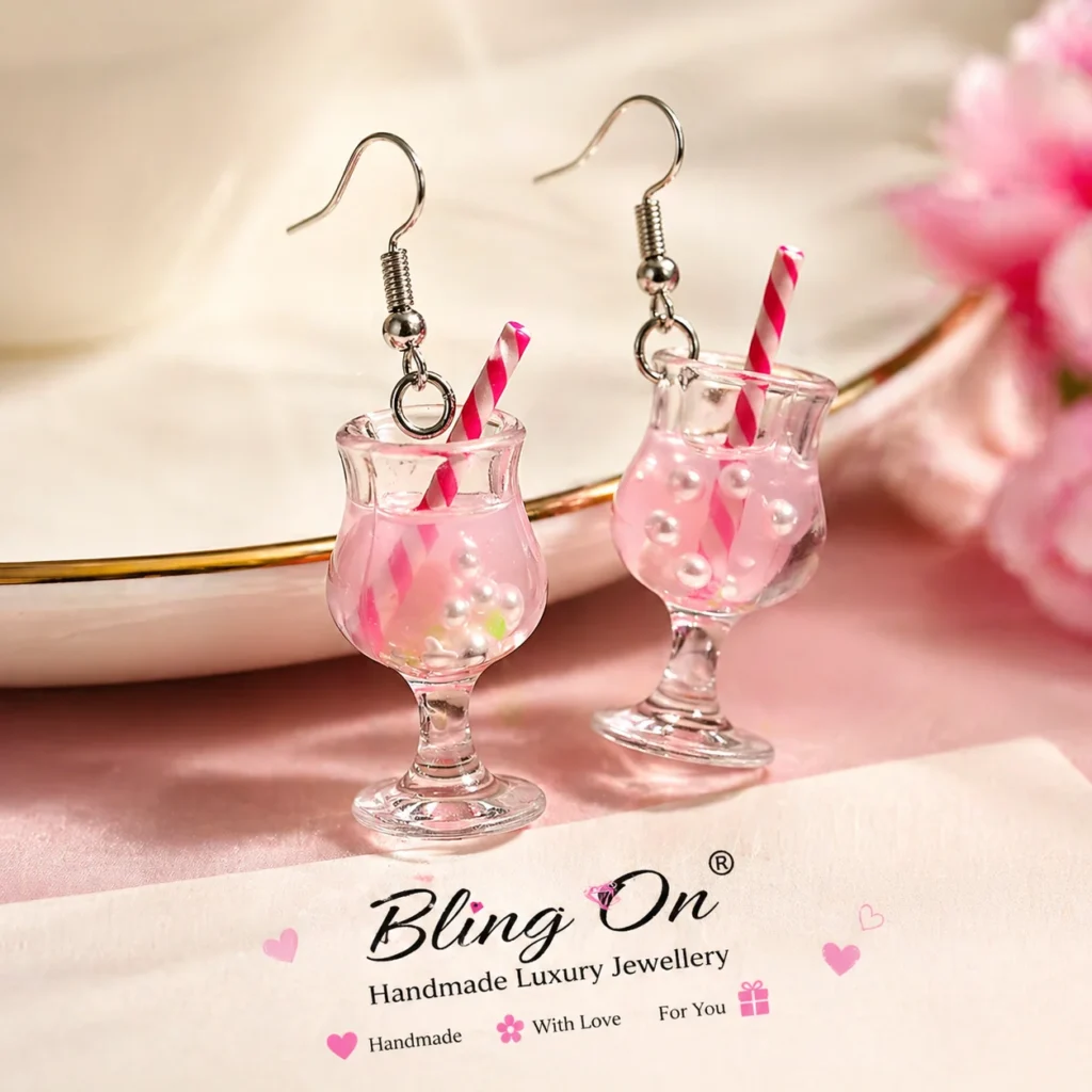 Pair of pink cocktail-glass earrings with striped pink straws and pearl beads on a pastel pink surface beside a Bling On jewelry card.