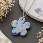 Silver Necklace Pendant Shaped Like a Four petal Flower with Blue and Teal Speckled Enamel Resting on a Dark Surface with Dried Floral Accents  Bling On