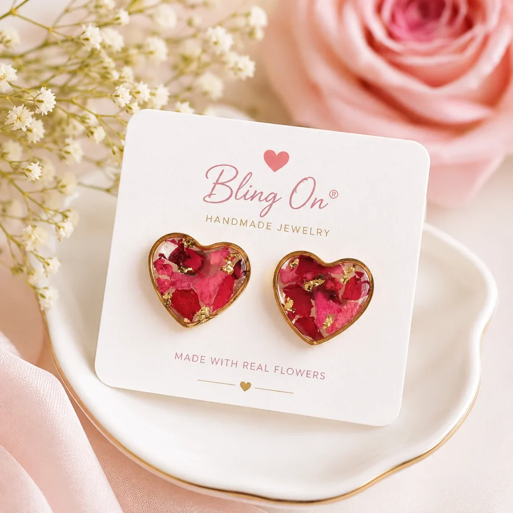 Heart-shaped pink resin earrings in gold settings on a Bling On card with 'Made with real flowers' text, placed on a plate with soft pink decor in the background.
