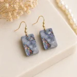 Rectangular Gray Resin Earrings with Iridescent Confetti Inlays on Gold Hooks Shown on a Light Stone Surface with Dried Flowers Nearby  Bling On