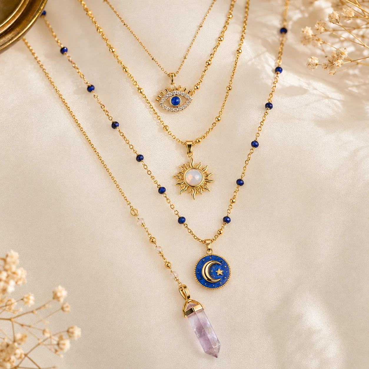Layered gold necklaces with blue beads and multiple pendants on cream satin backdrop, including an eye charm, sun pendant, moon and star pendant, and a lavender crystal point.
