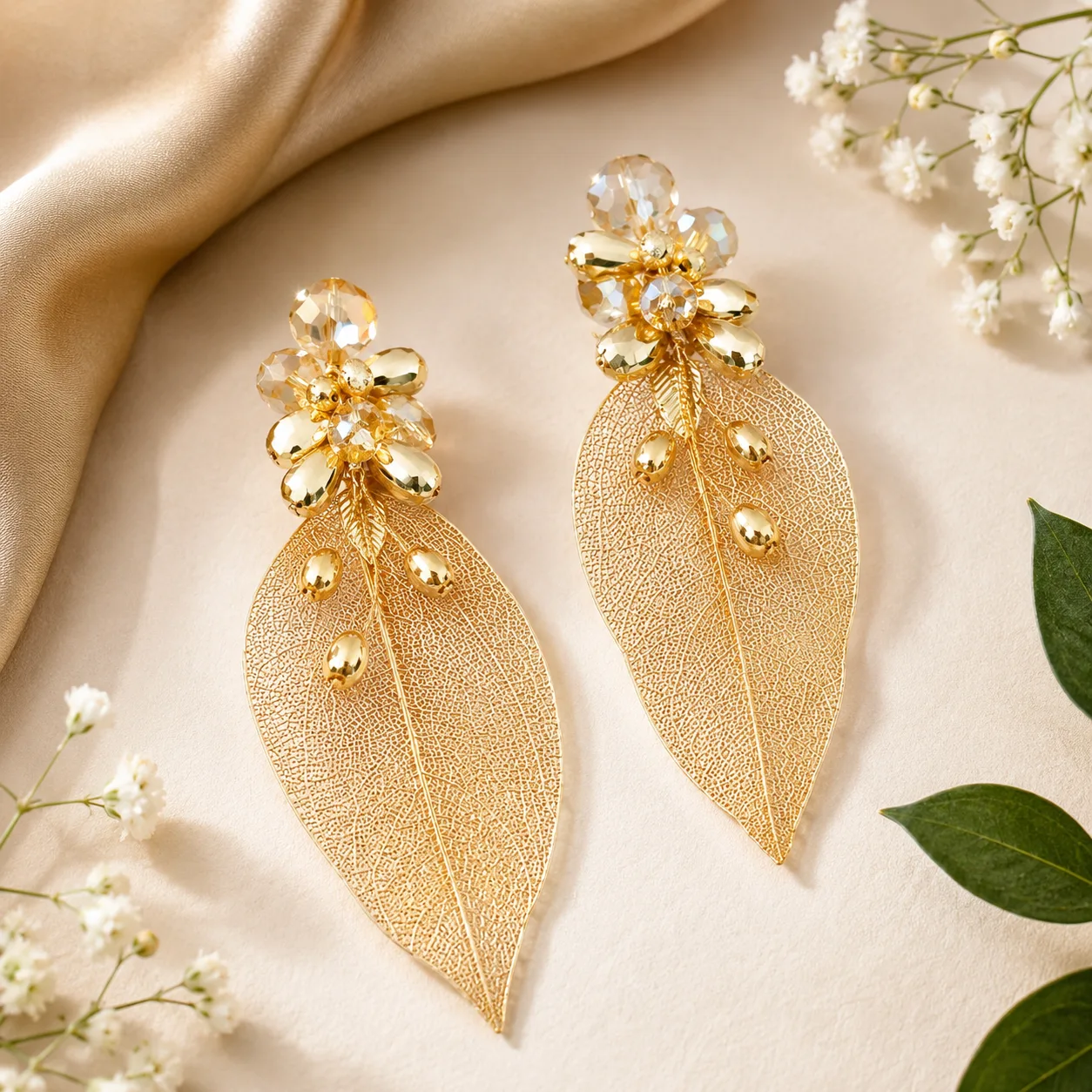 Gold-tone leaf earrings with crystal flower cluster at the top, on a beige fabric backdrop.