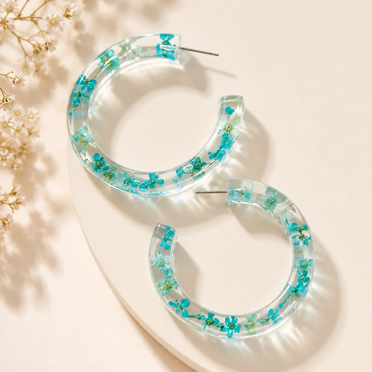 Clear resin hoop earrings embedded with blue flowers on a pale surface with dried blooms nearby.