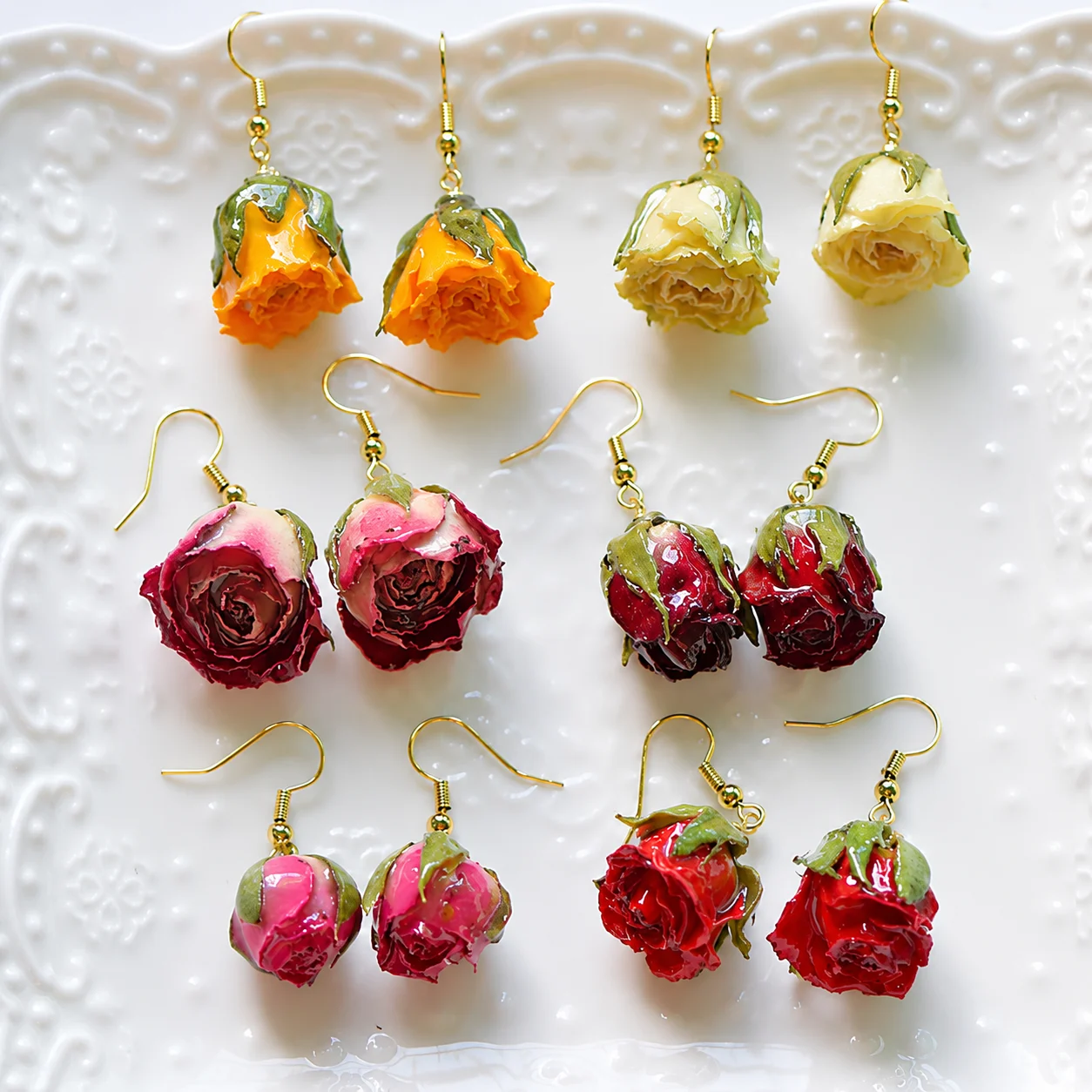 Dried rosebud chandelier earrings in orange, pink, and red hues on a white decorative plate with gold hooks and findings