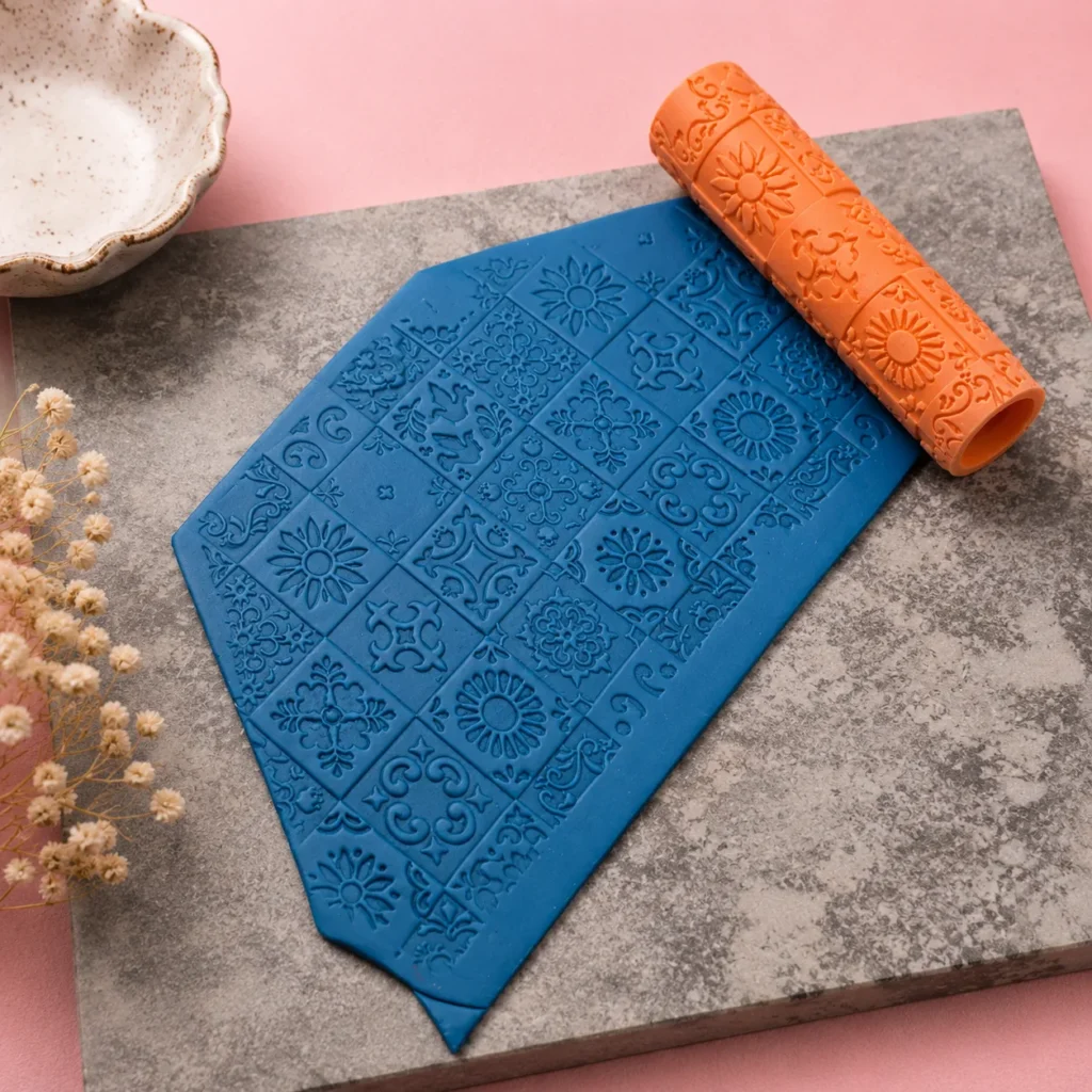 Blue embossed fondant embossing mat with a floral tile pattern and an orange patterned rolling pin on a gray surface, pink background.