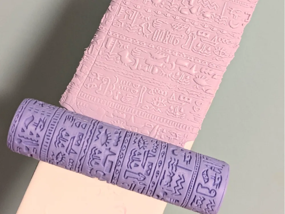 Pink embossing sheet and a purple fondant roller with Egyptian hieroglyph patterns on a light surface.