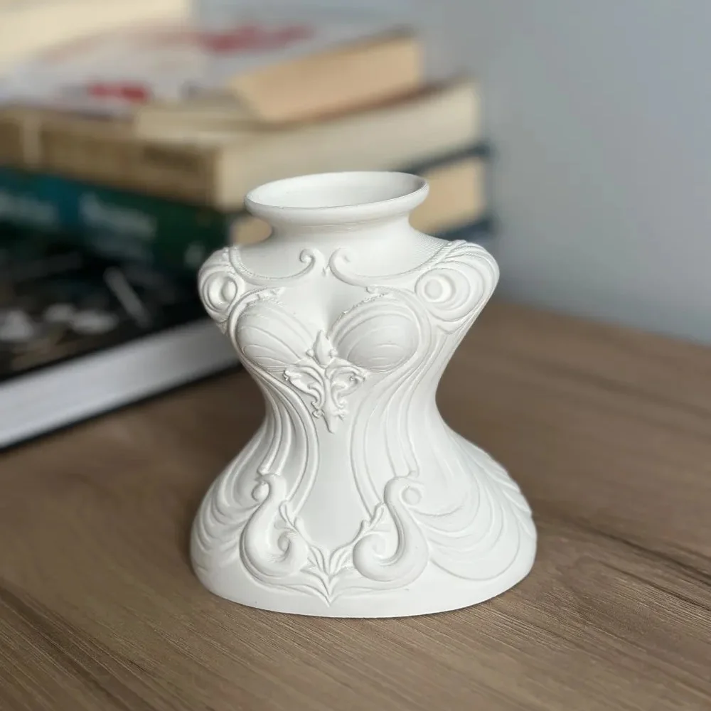 White decorative vase with raised scrollwork pattern sits on a wooden surface, blurred books in the background